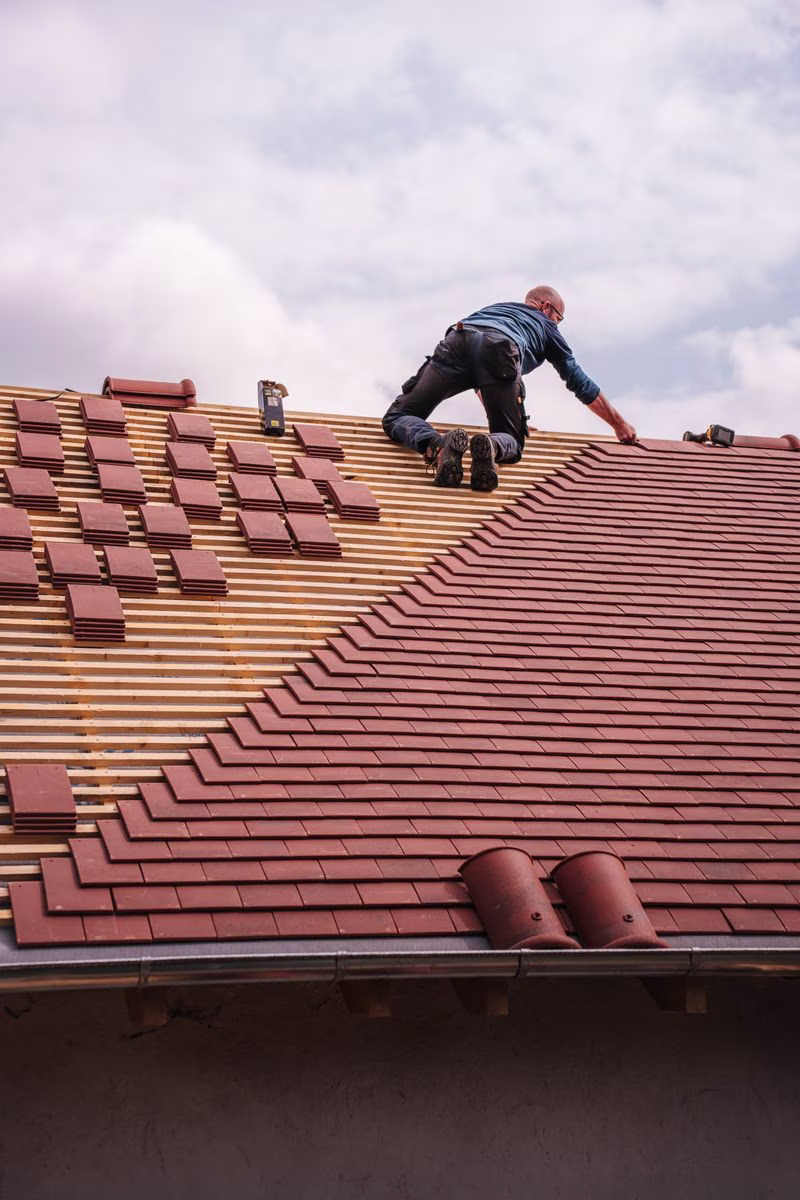 A roofer carefully installs shingles on a new residential roof under a cloudy sky.